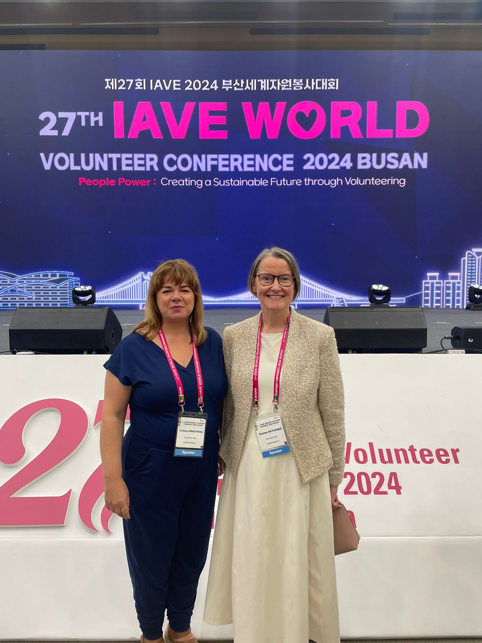 Lindsay & Denise standing in front of the IAVE World Volunteer Conference 2024 sign