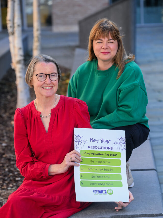 Two smiling women, one in a red dress, the other wiearing a green jumper holding a sign for New Years resolutions