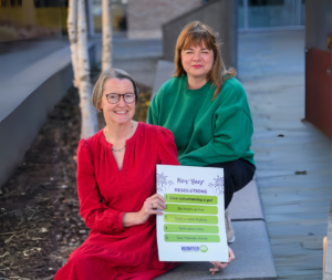 Two smiling women, one in a red dress, the other wiearing a green jumper holding a sign for New Years resolutions