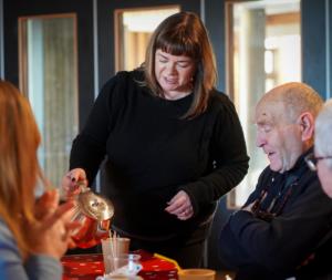 3 people sitting at a table while a woman in a black jumper pours tea from a teapot