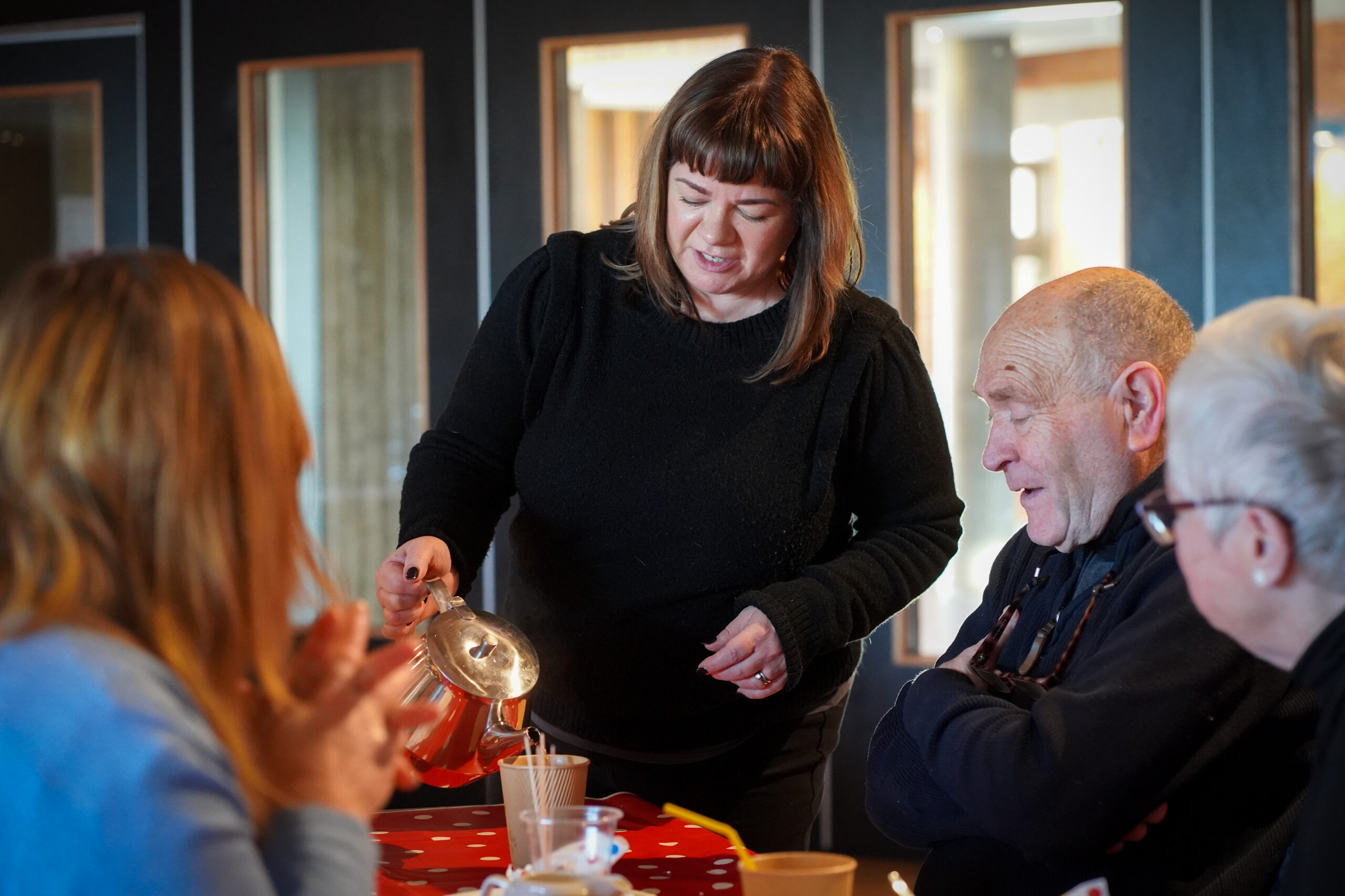 3 people sitting at a table while a woman in a black jumper pours tea from a teapot